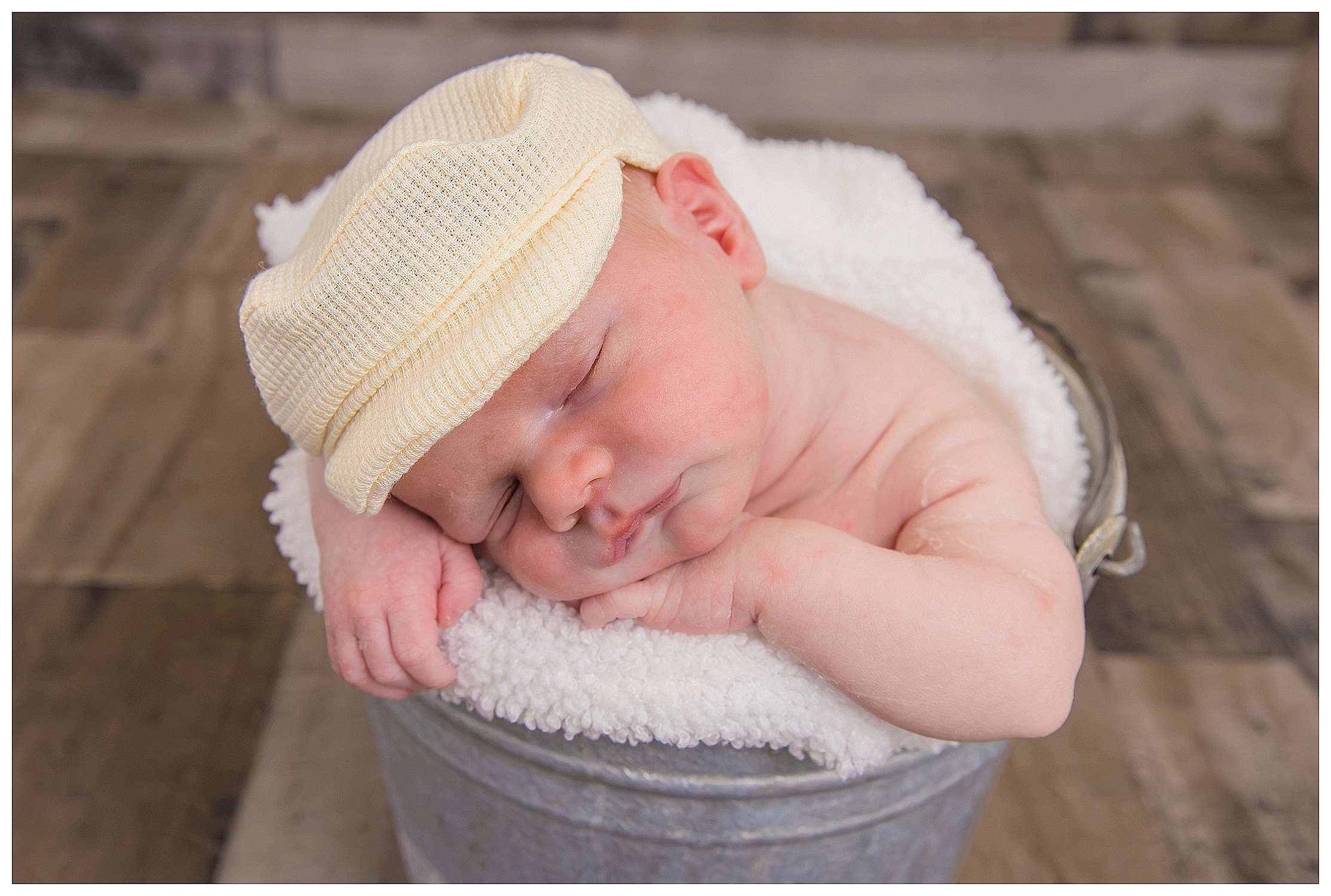 Sleeping newborn baby wearing a soft knit cap, curled up in a cozy blanket inside a small metal bucket, captured during a posed studio session.
