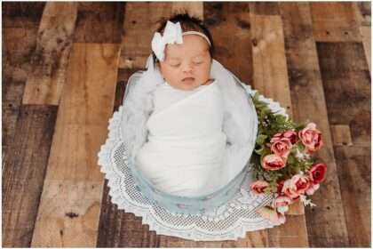 Newborn baby girl wrapped in white fabric during a Downtown Everett Studio newborn session, posed inside a floral keepsake box with soft pink flowers nearby.