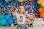 Three-year-old girl smiling during a colorful Lisa Frank inspired jungle birthday photoshoot with rainbow balloons and cartoon jungle animals in an Everett photography studio.