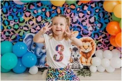 Three-year-old girl smiling during a colorful Lisa Frank inspired jungle birthday photoshoot with rainbow balloons and cartoon jungle animals in an Everett photography studio.