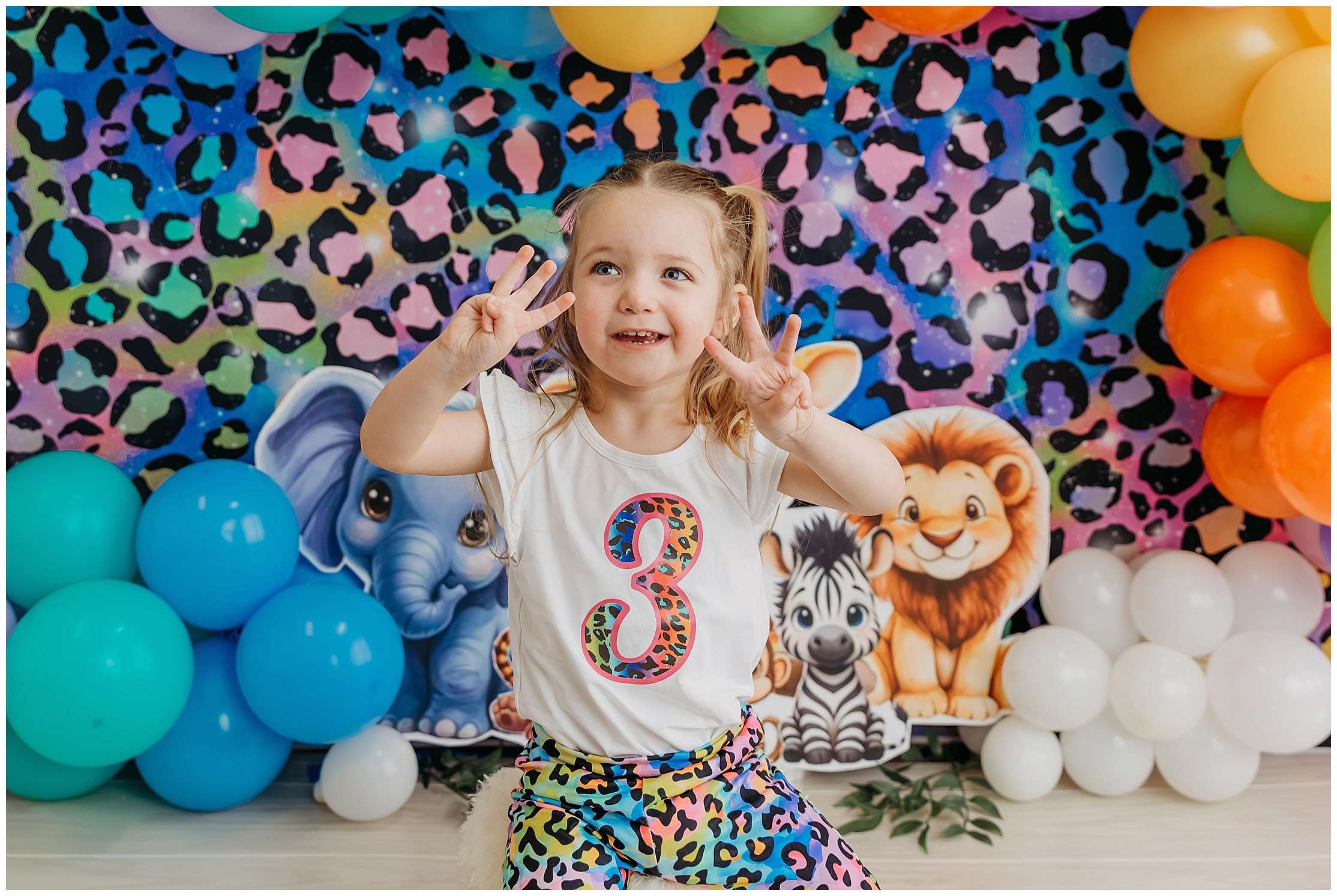 Three-year-old girl smiling during a colorful Lisa Frank inspired jungle birthday photoshoot with rainbow balloons and cartoon jungle animals in an Everett photography studio.