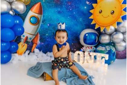 Baby boy celebrating his first birthday with a space-themed cake smash setup featuring rockets, planets, balloons, astronaut décor, and a glowing ONE sign.