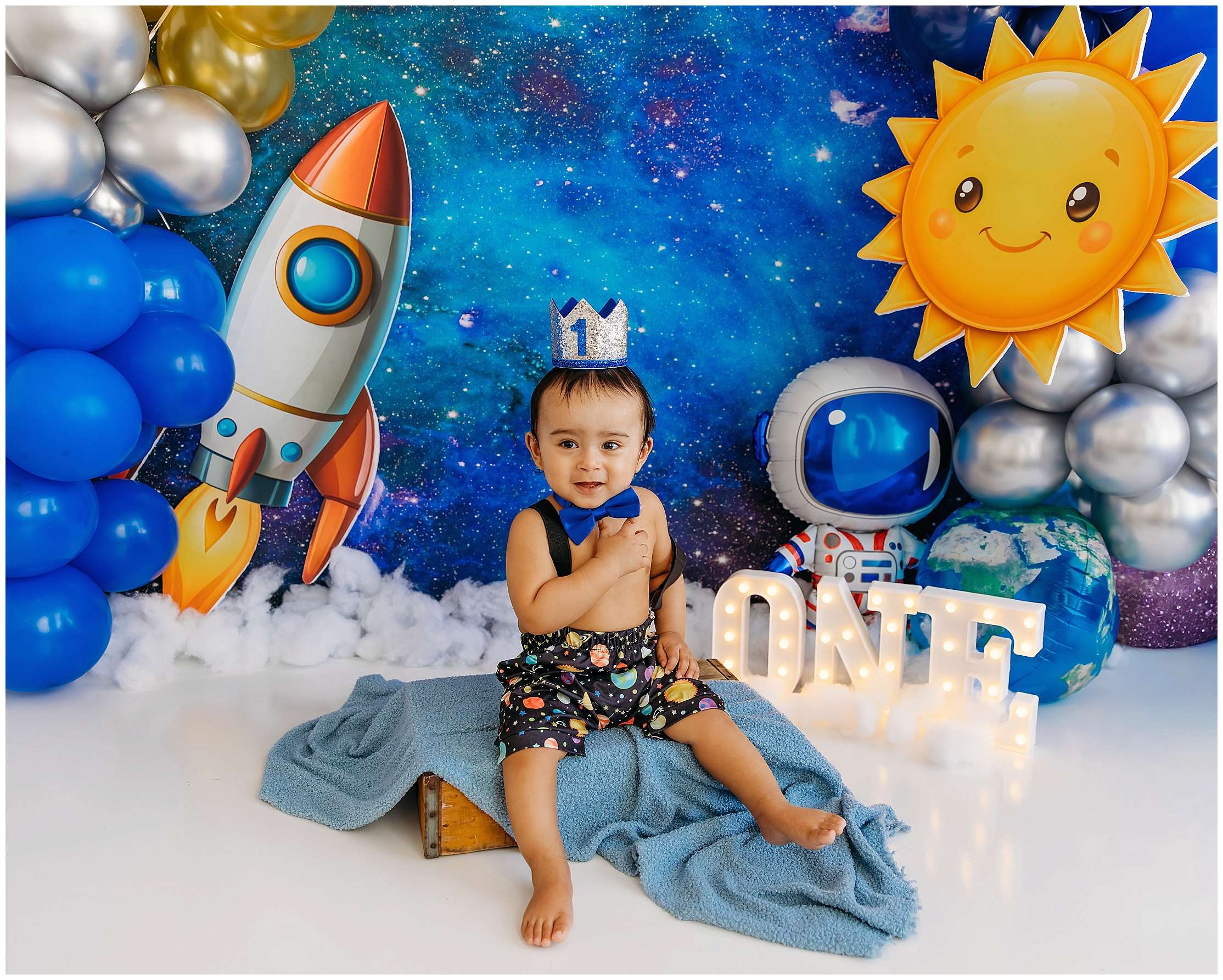 Baby boy celebrating his first birthday with a space-themed cake smash setup featuring rockets, planets, balloons, astronaut décor, and a glowing ONE sign.